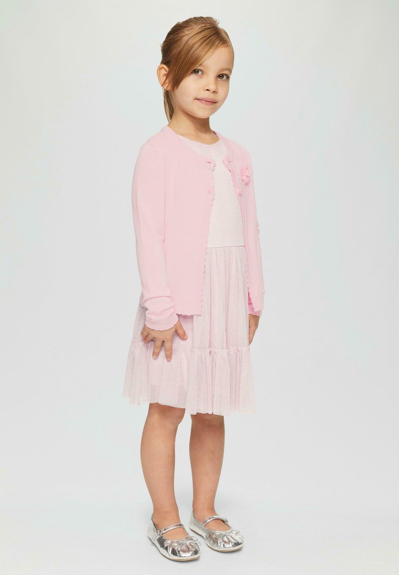 Lichtroze cardigan met knoopsluiting en bloemenaccent, laagjes witte jurk met geplooide rok, en glanzende zilveren ballerina's.