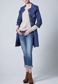 Granatowy trench z dużymi guzikami, kremowy sweter dziany, niebieskie dżinsy i brązowe botki za kostkę z grubym obcasem. Zrolowane mankiety dżinsów.