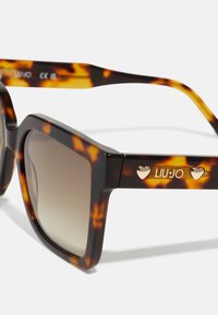 Ochelari de soare tartan cu ramă pătrată, lentile cu gradient și accente dorite în formă de inimă din aur pe brate. Logo-ul mărcii gravat pe o parte.
