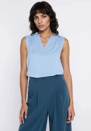 Kobieta ubrana w niebieską, bezrękawną bluzkę z dekoltem w serek oraz szerokie, wysokie spodnie w kolorze morskim, stojąca na białym tle.