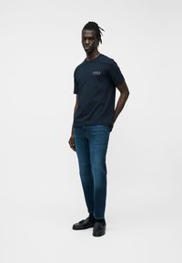 Fiatal férfi áll, kezeit a háta mögött tartva, fekete Tommy Hilfiger pólót, kék farmert és fekete mokaszint viselve, egyszerű fehér háttér előtt.