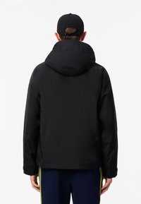 Giacca nera impermeabile con un grande cappuccio regolabile, caratterizzata da una texture liscia e senza hardware visibile. Indossata sopra pantaloni blu scuro.