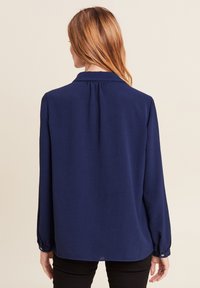 Blusa lunga blu navy con polsini a bottone, colletto, pieghe sul retro e una texture liscia. Abbinata a pantaloni neri.
