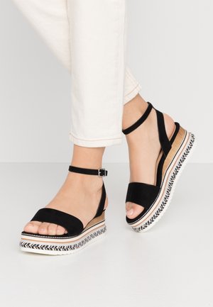 Sandales compensées à plateforme noire avec brides à la cheville, portées avec un pantalon blanc, montrant les pieds et une partie des jambes contre un fond uni.