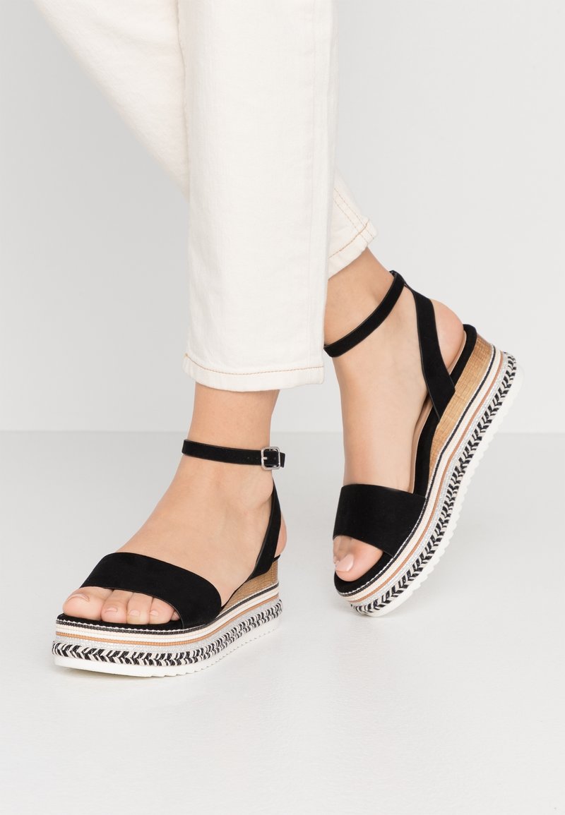 Sandales compensées à plateforme noire avec brides à la cheville, portées avec un pantalon blanc, montrant les pieds et une partie des jambes contre un fond uni.