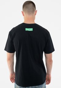 Black cotton t-shirt with a crew neck and short sleeves, featuring a square green logo patch on the upper back.
