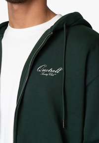 Dunkelgrüner Reißverschluss-Hoodie aus Baumwollmischung mit einem weißen gestickten Logo auf der Brust und einer Kordelzugkapuze. Darunter ein weißes T-Shirt.