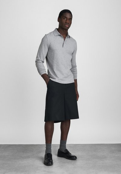 Sivý polo s dlhým rukávom a golierom, skombinovaný s čiernymi kraťasmi po kolená. Model má na sebe sivé ponožky a čierne elegantné topánky. Jednoduchý, moderný dizajn.