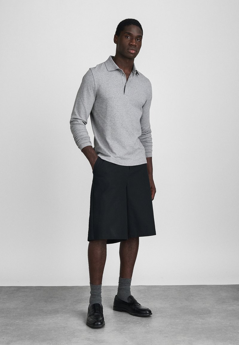 Polo grigio a maniche lunghe con colletto, abbinato a pantaloni corti neri fino al ginocchio. Il modello indossa calze grigie e scarpe eleganti nere. Design semplice e moderno.
