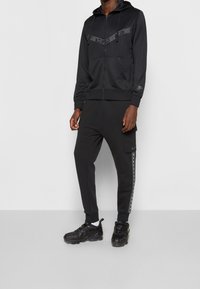 Černá mikina na zip a tepláky, s hladkou texturou, s logem Nike na hrudi a bočním pruhem na kalhotách. Černé tenisky.