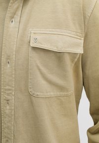 Beige Langarmshirt mit einer einzelnen Brusttasche und gesticktem Detail, aus weichem Stoff mit leicht strukturiertem Finish.
