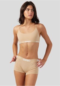 Beige sports-bh med tynne stropper og et hvitt logobånd, sammen med matchende beige shortser med en hvit logolett.