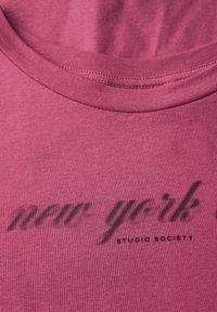 Detailní záběr na výstřih růžového trička, na kterém jsou černým kurzívou s malými písmeny vytištěny slova "new york studio society".