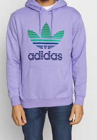 Lila bomullshoodie med en framficka. Har en gradient i grönt och blått randigt Adidas-logotyp på bröstet. Avslappnad passform med dragsnodd i huven.