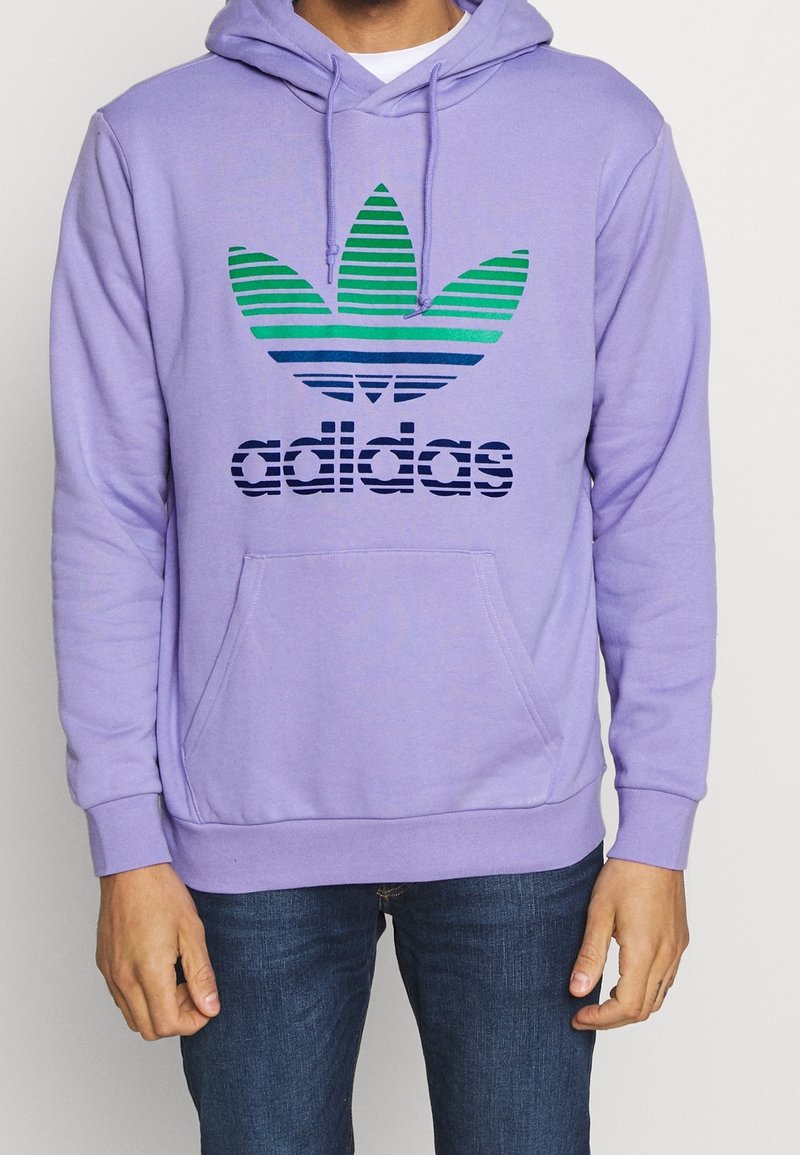 Lila bomullshoodie med en framficka. Har en gradient i grönt och blått randigt Adidas-logotyp på bröstet. Avslappnad passform med dragsnodd i huven.