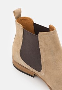 Bottine Chelsea en daim beige avec panneau latéral élastique foncé, intérieur en cuir, languette à l'arrière, et semelle en bois empilée avec semelle extérieure en caoutchouc noir.