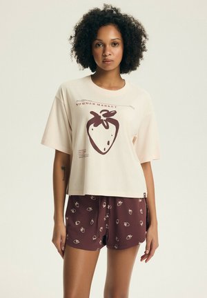 Kobieta z kręconymi włosami, ubrana w beżowy t-shirt z dużym nadrukiem truskawki oraz bordowe spodenki z małymi nadrukami truskawek.