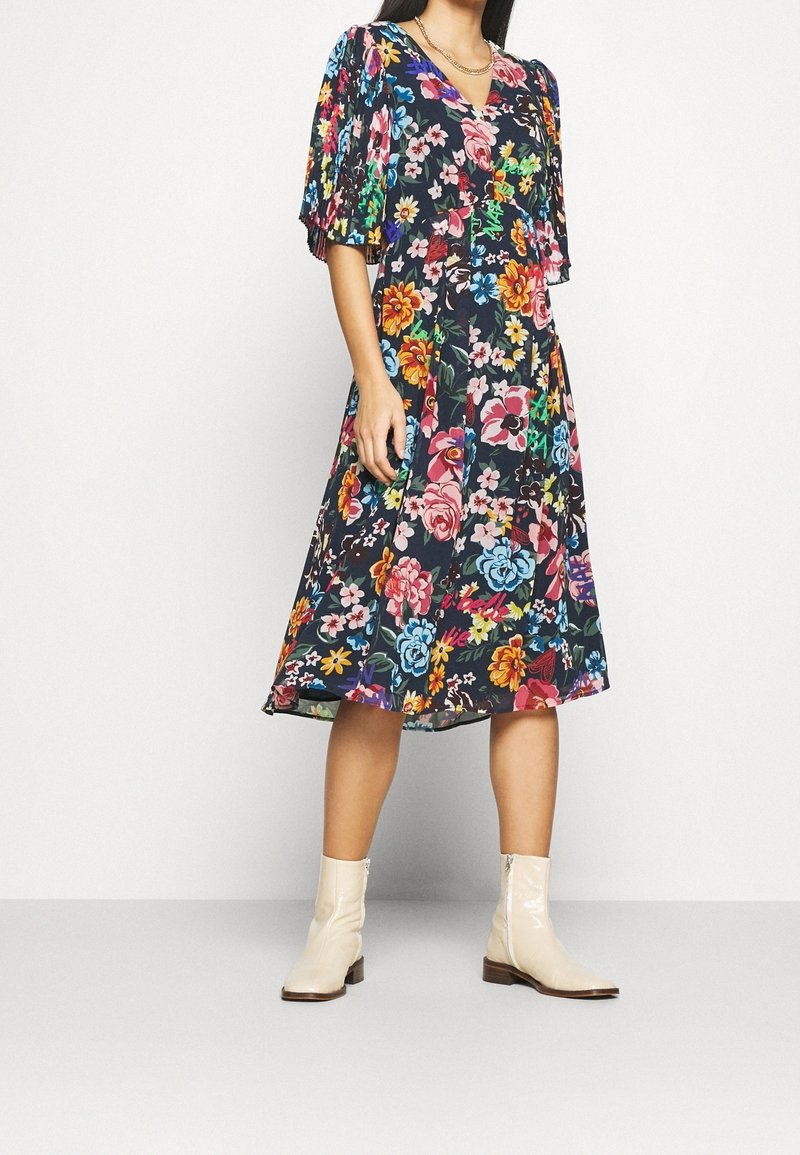 Femme portant une robe fleurie foncée longueur genoux avec des manches courtes et larges et des bottines beiges, debout devant un fond blanc uni.