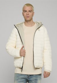 Hvit dunjakke med tofarget, quiltet design; har en grå hette og svarte glidelåsdetaljer. Brukes over en beige skjorte.