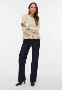 Krämfärgad stickad tröja med volangkant, med ett strukturerat mönster och knäppning fram, kombinerad med mörkblå vida jeans.