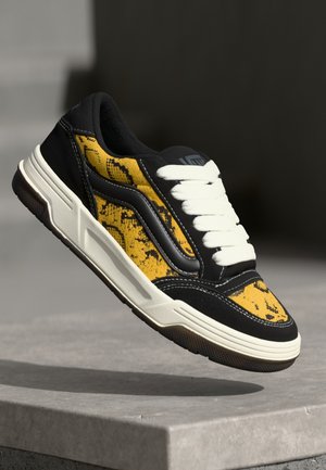 Schwarzer und gelber Sneaker mit weißen Schnürsenkeln, der über einer Steinoberfläche vor dunklem Hintergrund schwebt.