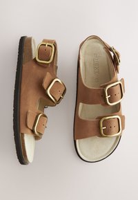 Sandalias de ante marrón con tres hebillas doradas, diseño de punta abierta y plantilla texturizada. Suelas de goma que proporcionan agarre y soporte.