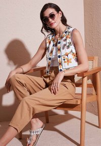 Embroidered white sleeveless vest with floral motifs, paired with beige pants. The model wears sunglasses and decorative sandals. Wood chair background.
