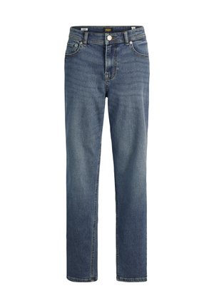 Personne portant un t-shirt blanc uni à manches courtes, un jean bleu et des baskets noires et blanches, debout devant un fond uni.