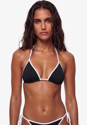 Nainen, jolla on märät hiukset, musta bikini valkoisella reunuksella, seisoo yksinkertaisen valkoisen taustan edessä ja katsoo eteenpäin neutraalilla ilmeellä.