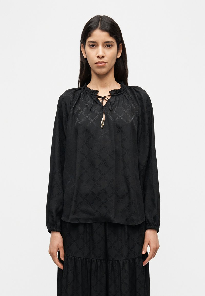 Μαύρη μπλούζα με διακριτικό υφασμάτινο μοτίβο, συγκεντρωμένη λαιμόκοψη με κορδόνι, μακριά μανίκια και άνετη εφαρμογή.