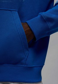 Niebieska bluza z kapturem z przednią kieszenią, ściągaczami na mankietach i u dołu, o gładkiej fakturze i przeszywanych szwach.