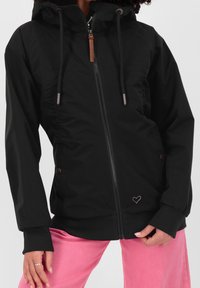 Donna che indossa una felpa nera con zip e coulisse e pantaloni rosa, con le mani rilassate sui fianchi, su uno sfondo semplice.