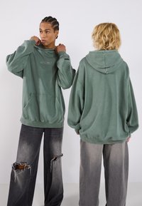 Hoodie oversize verde oliva con tasca frontale e cappuccio con lacci, che presenta il logo "URTRN" sul petto. Indossato con jeans grigi strappati.