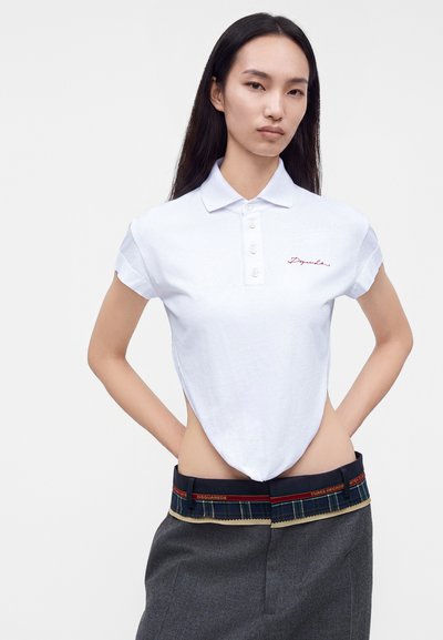 Femme aux longs cheveux foncés portant un polo blanc court et un pantalon gris avec une ceinture sombre à motif partiellement visible.