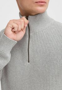 Suéter gris con cuello cerrado y cremallera, que presenta una textura acanalada. El tejido parece suave y cómodo.
