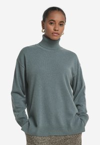 Kvinna i turkos stickad polotröja och guldörhängen, med händerna i fickorna, stående mot en neutral vit bakgrund.