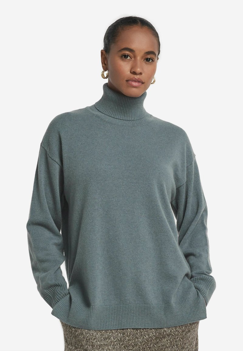 Kvinna i turkos stickad polotröja och guldörhängen, med händerna i fickorna, stående mot en neutral vit bakgrund.