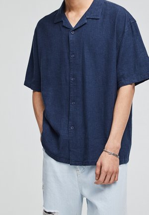 Homme portant une chemise bleu foncé à manches courtes boutonnée et un short en jean bleu clair déchiré, avec un bracelet en argent au poignet gauche.