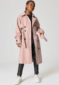 Ljusrosa trenchcoat med stora svarta knappar, bälte i midjan och vida ärmar. Matchas med svarta leggings och vita sneakers.