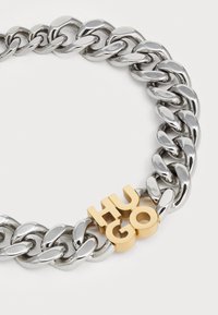 Bracciale in argento metallico con catena a maglie spesse e lettering "HUGO" in oro come dettaglio distintivo. Superficie liscia con finitura lucida.