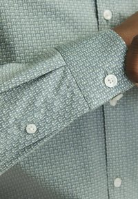 Camicia a maniche lunghe verde con motivo, dotata di bottoni bianchi, caratterizzata da un tessuto dalla texture particolare e un design dei polsini aderente. Il motivo è composto da piccole forme geometriche.