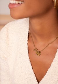 Collier doré avec des pendentifs en forme de cœur double ; chaîne texturée et fermoir à bascule ; porté sur un vêtement doux de couleur crème.
