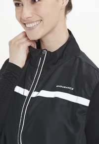 Svart jacka med dragkedja framtill, hög krage och vita horisontella ränder. Materialet verkar lätt med texturerade ärmsektioner.