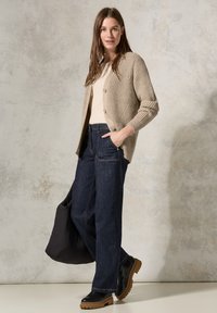 Beige ribbed cardigan dengan kancing, atasan ringan, celana jeans lebar gelap, dan sepatu boots chunky sole hitam. Model memegang tas puffer hitam.