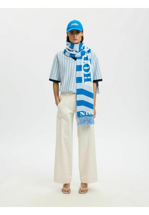 Vrouw met een blauwe pet, gestreept overhemd, witte wijde broekspijpen, blauw-witte sjaal en beige sandalen, staand tegen een witte achtergrond.