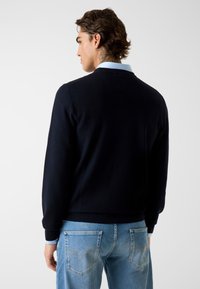 Marineblauwe gebreide trui met een geribde zoom, gecombineerd met een lichtblauw overhemd met kraag eronder en een ontspannen pasvorm jeans met zichtbare stiksels.