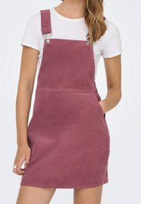 Femme portant une robe chasuble en velours côtelé rose poussiéreux sans manches par-dessus un t-shirt blanc à manches courtes, avec une main dans la poche de la robe.