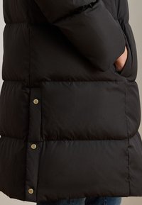 Juodas pūkinis švarkas su siūtu raštu, lygia tekstūra ir auksinių detalių užsegimuose bei kišenėse.