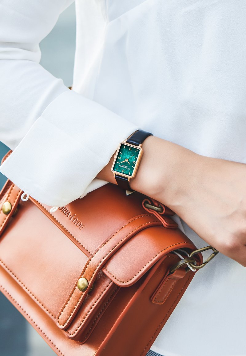 Gul rektangulär klocka med grön urtavla och svart läderarmband på en handled, bredvid en brun läderhandväska med mässingsdetaljer.