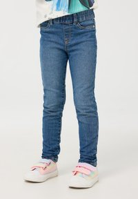 Niebieskie dżinsy skinny z klasycznym stanem, noszone z pastelowymi trampkami w tęczowych kolorach, z rzepami i teksturowaną cholewką.